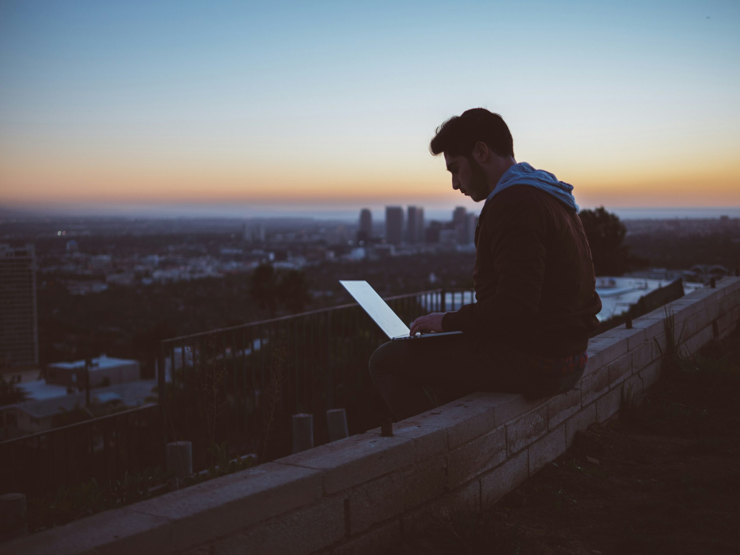 Guy sitting with a laptop on his knees, overlooking a city valley at night.